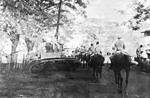 A long column of men on horseback moving down a road. A tank is parked beside the road.