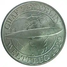 A circular silver coin, with a raised edge. Inside this is written, in raised capitals, "Graf Zeppelin/Weltflug 1929". The two halves of the inscription are separated by a depiction of the airship flying from left to right, against a background showing the northern hemisphere of the world marked with stylised lines of latitude and longitude. At the bottom of this is a letter "A".