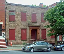 A two-story brick facade with red boards in the windows and brownstone around the entrance. Parked in front of a tree on the street is a dark gray late-2000s Honda Civic