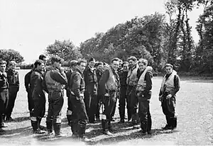 Black-and-white photo of a group of young men wearing military uniforms and life preservers standing in a field edged with trees