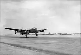 Black-and-white photo of a four engined World War II-era monoplane aircraft flying just above the ground in front of an open area