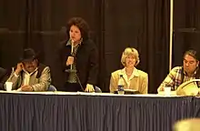 Landscape photograph displaying woman with microphone speaking and 3 other individuals sitting next to her. Susan Masten, standing, with Secretary Gale Norton (right of Masten) and Three Affiliated Tribes Chairman Tex Hall (right of Norton)