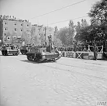 Men of the 51st (Highland) Infantry Division, mounted in Universal Carriers, drive past a podium of senior officers during a victory parade.
