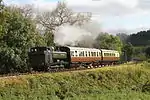 A green pannier tank locomotive is travelling from right to left, bunker first, along a single track on a grassy embankment. The locomotive is pulling two passenger carriages painted cream and brown, and a plume of smoke is passing over the first carriage. In the background are a number of green trees, but behind them are some taller trees that have lost most of their leaves.