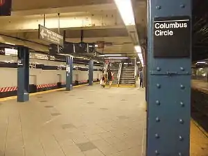 Uptown platform of the IND station. There are tracks on either side of the platform. To the right is a blue column containing a sign with the words "Columbus Circle". In the background is a staircase.
