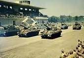Colour picture of Sherman tanks passing a saluting diaz in an autodrome.