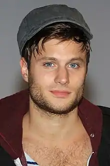 A man with blue eyes and a flatcap faces the camera. He wears a navy and white stripped top with an unzipped hoodie over the top and his chest hair on show