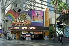 Ground-level view of the front entrance to a casino; a very prominent, colorful sign features a rainbow and a pot-of-gold. There are slot machines inside the building, and glass windows and part of a skyscraper are visible above the sign.