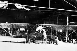 Front view of single-engined biplane and two men in front of a large open hangar