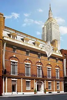 A multi-leveled brick building with many whitepaned windows with skycraper visible in the top right corner