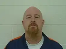Photo of inmate John Eric Armstrong wearing standard uniform of inmates in the Michigan Department of Corrections consisting of a white undershirt and a dark blue button up shirt with an orange stripe on each shoulder