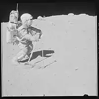 John Young using the rake tool to collect a sample (67700) at the southeast rim, with the rover at the central horizon, and white breccia boulders at the right horizon