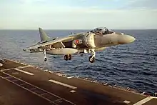 Grey jet aircraft executing a vertical takeoff from aircraft carrier at sea. Under each of the angled-down wings is an external fuel tank.