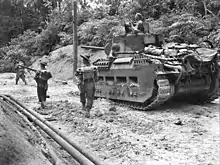 An armoured vehicle moves along a sandy road surrounded by jungle. Infantrymen advance alongside