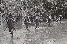 Soldiers wearing slouch hats and shorts wade along a watercourse