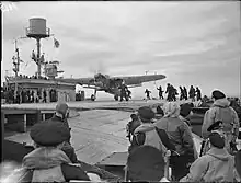 Black and white photograph of a single-engined monoplane on the flight deck of a World War II-era aircraft carrier. Large numbers of men are watching the aircraft or running towards it.