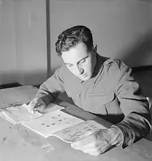 A man sits at a table with a booklet and paper. There are no identifying marks on his uniform.
