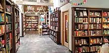 A welcoming room of bookshelves. Multiple rainbow decorations are visible, and a queer fiction section is prominently displayed.