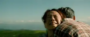 Atop a mountain on a sunny day, a man hugs his wife as the wife cries hopelessly