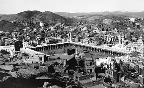 An old photograph showing a black, cubic structure enclosed by rectangular arcade surrounded by buildings and hills