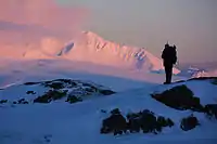 A hiker standing on a rocky knoll, the pink early morning light illuminating snow-covered Mt. Moffett in the background