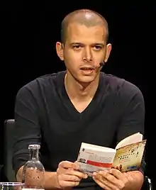 front view of a male seated at a table reading from a book