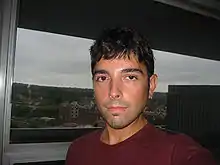 Dark-haired man with unkempt hair. He is wearing a red shirt. A silver chain around his neck and an earring in his left ear can be seen.