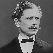 Monochrome photograph portrait of a man in his mid-20s shown from the shoulders up, wearing a dark coat, white collared shirt and dark, thin, bow tie, the man's shoulders squared forward but his head turned somewhat to the right, looking right, with light-colored hair in short, oiled waves on the head, light-colored eyebrows, and a wide, light-colored mustache extending just beyond the corners of the mouth.