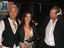 A man with blonde hair wearing a white T-shirt, a smiling woman with brown hair wearing a dress and another man wearing a suit and touching his head.
