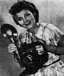 Black and white photograph of a woman in a printed dress holding a large press camera with a flashbulb attachment in front of her