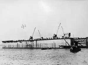 A photo of a rectangular structure with cranes mounted on it floating on a calm body of water. Several boats are visible in front of the structure