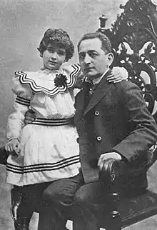 A white man with his daughter, about ten years old, seated on his lap; she is wearing a white dress with a wide collar and black striped embellisments; he is wearing a suit