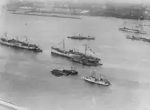 An overhead photograph of eight ships. At the bottom of the photograph is one side of a harbor with the ships in the water. Five ships are displaying decorative bunting. At the top of the photograph is the opposite side of the harbor.