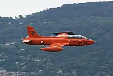 A military jet in flight, painted bright orange with Italian Air Force roundels, against the backdrop of Lake Maggiore mountains in Italy