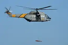 Spanish Air Force Aérospatiale SA330J Puma of 801 Squadron flying in an airshow. It is lifting a stretcher with a hoist. On the side of the helicopter is lettering reading "SAR", in yellow against the military grey color scheme.