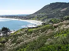 a sweeping beach with blue sea on the left and green hills on the right, small wooden dwellings dotted on the hills