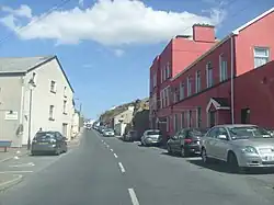 A road between two buildings, one red and the other pebbledash, with cars parked on either side.