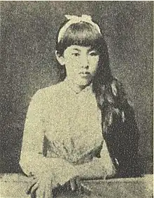 Grainy black and white photograph of a woman looking to the viewer's right. She has black hair and is wearing light-colored clothing. Her arms are folded in front of her on a raised surface like a table. Her hair is tied behind her ears with a light-colored ribbon with a small bow at the top of her head.