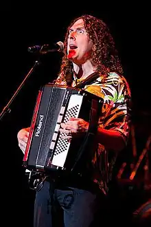 A photograph of "Weird Al" Yankovic, singing through a microphone and playing a harmonica.