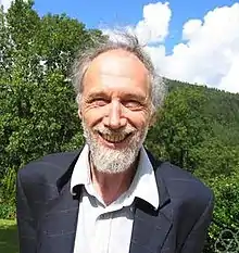 Photo of the upper body of Alain Connes with vegetation, blue sky, and clouds in the background