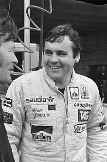 Alan Jones in a racing outfit smiling at another man