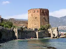 A eight-sided medieval tower built of red and yellow brick rises above a green sea in which swimmers play.  Stone walls run along the shore and further up from the tower.