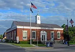 A low brick building with a square cupola in the middle of its metal roof. Wording on the front says "UNITED STATES POST OFFICE" and "ALBION NEW YORK". There is an American flag flying from a flagpole in front and hanging from a lamppost on the right.