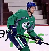 A Caucasian ice hockey player skating with gripping a hockey stick with both hands. He wears a green jersey with white and blue trim and a blue, visored helmet.