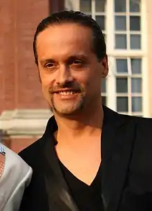 close-up of Alex Christensen wearing a black v-neck t-shirt and a black jacket, grinning and looking right of camera