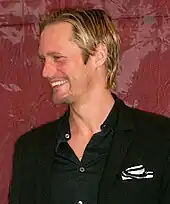 A man with blond hair and dressed in a black shirt and blazer smiles to his right