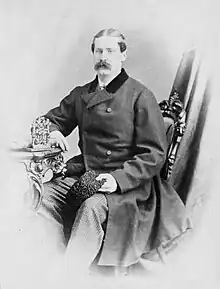 Fair-skinned man with dark hair and mustache, seated at table for a formal picture, wearing a mid-Victorian frock coat and holding a fur cap in his hand
