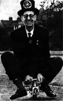 A smiling man with glasses wearing a dark suit and a hat riding a miniature bicycle