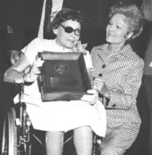 A white woman seated in a wheelchair, with an eyepatch, glasses, wearing a corsage; her legs are both prosthetics; she is holding a plaque; kneeling next to her is another white woman, First Lady Pat Nixon, wearing a checked suit and smiling
