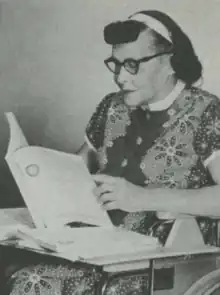 A white woman with dark hair, wearing a white headband, glasses and a patterned dress with short sleeves and a button front; she is seated in a wheelchair and reading a large softcover book